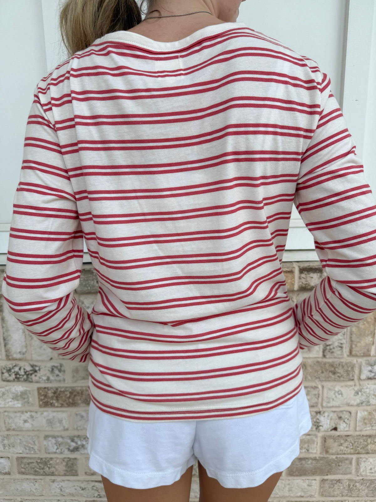 A person shown from the back wearing a long-sleeve white top with thin red horizontal stripes and white shorts, standing against a light brick wall.