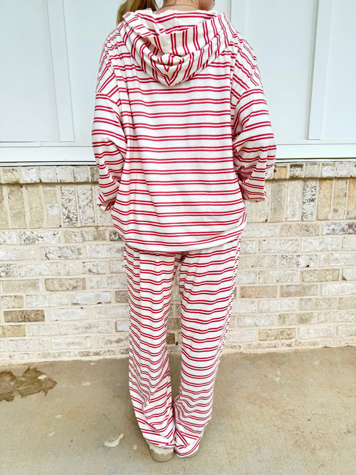 A person shown from the back wearing a matching red and white striped hooded sweatshirt and sweatpants set, standing outdoors against a light brick wall.