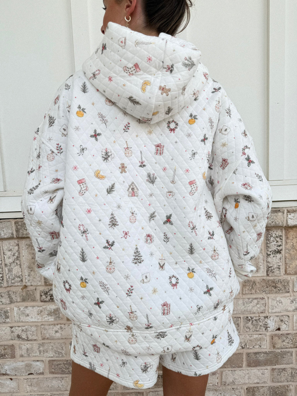 A person shown from the back wearing a quilted white hooded sweatshirt and matching shorts with a small multicolor holiday print, standing against a white paneled wall and brick base.