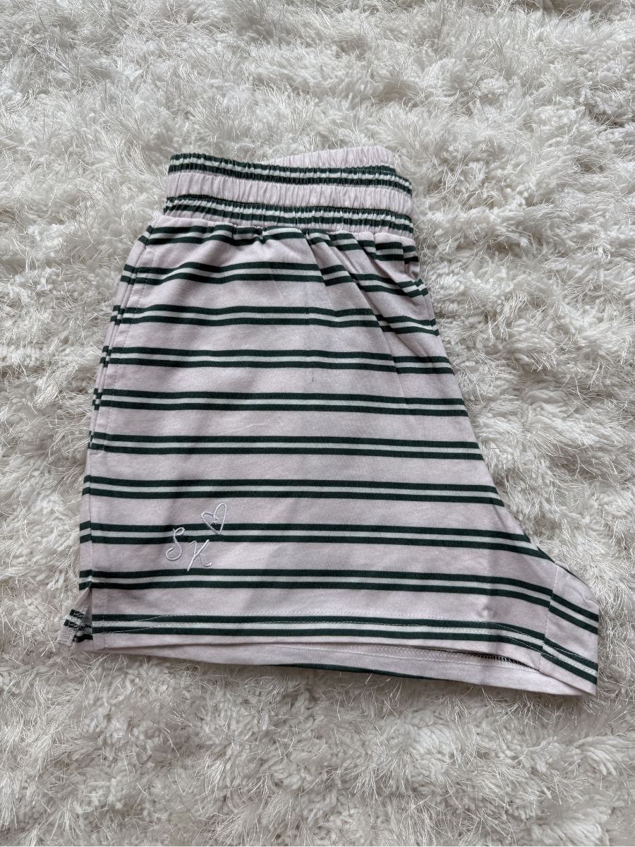 A children's skirt with horizontal dark green stripes on a light pinkish-beige base, featuring an elastic waistband and a small white embroidered logo near the lower left hem, laid flat on a white shag rug.