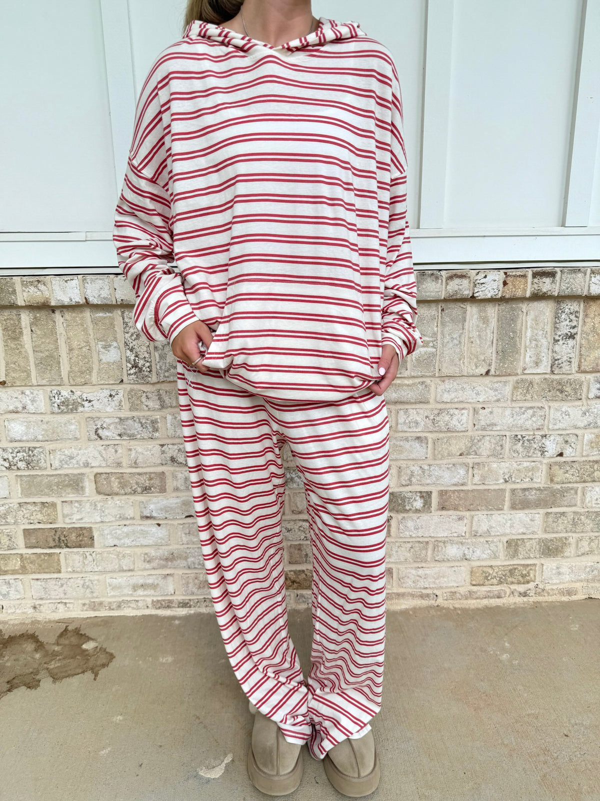 A person wearing a matching red and white striped lounge set with a hooded sweatshirt and sweatpants, standing in front of a light brick wall.