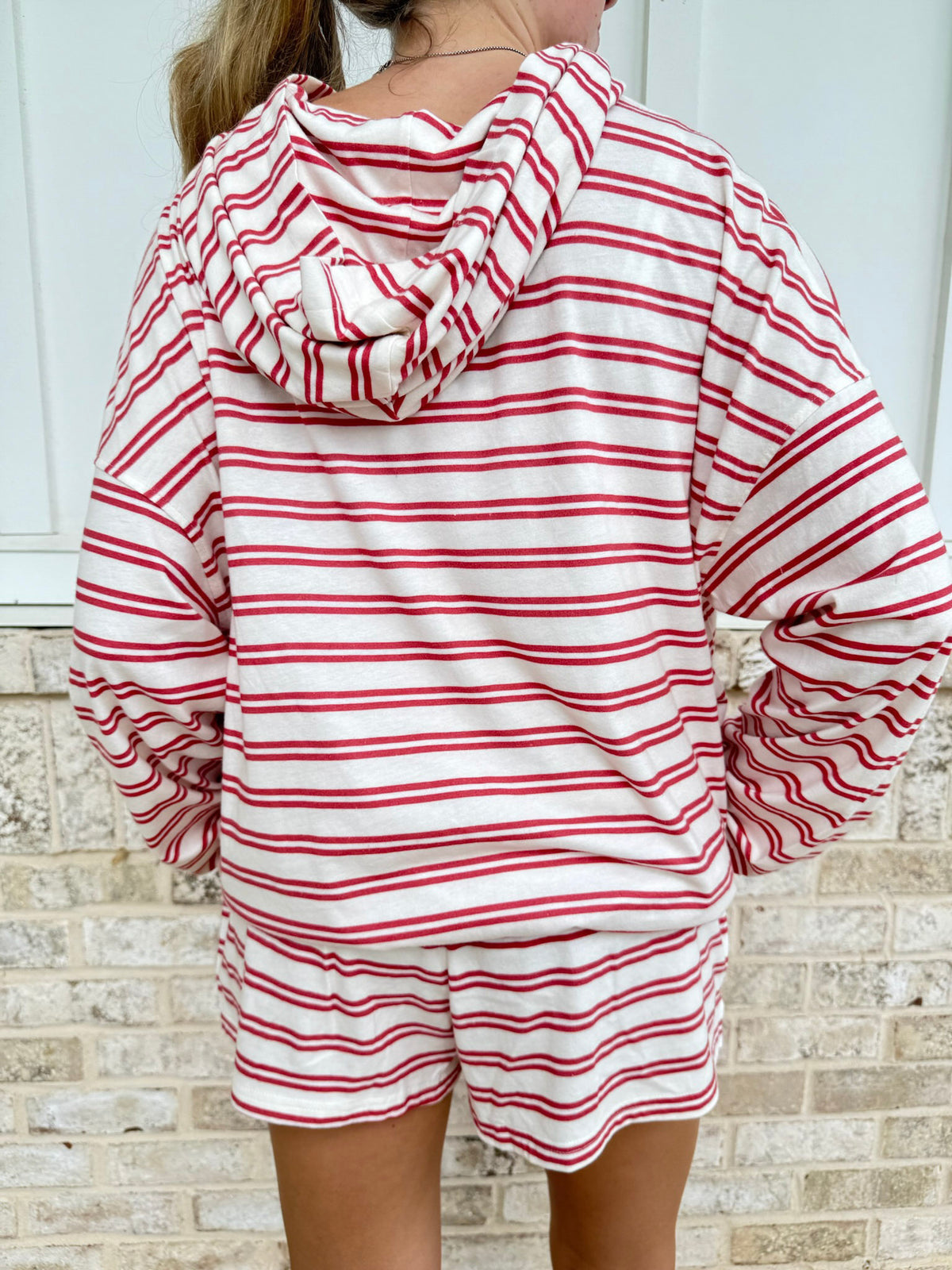 A person shown from the back wearing a red and white horizontally striped hooded sweatshirt and matching shorts, standing against a light brick wall.