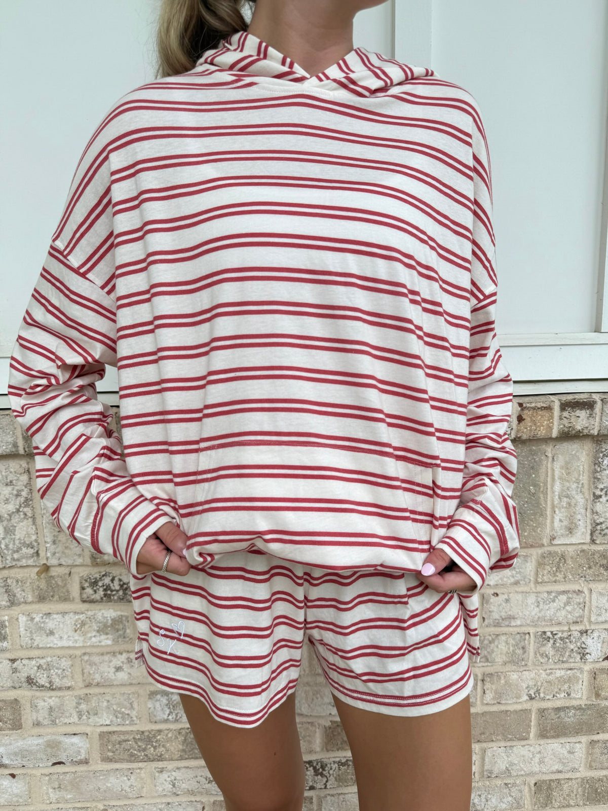 A person wearing a matching red and white striped lounge set, featuring a long-sleeve hooded sweatshirt and coordinating shorts, standing against a light brick wall.