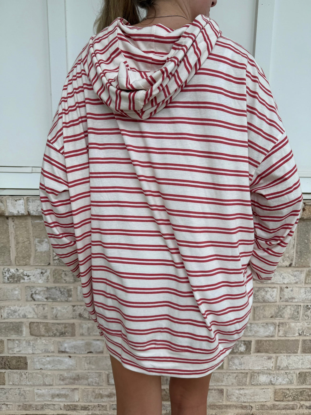 A person shown from the back wearing an oversized hooded sweatshirt with horizontal red and off-white stripes, standing in front of a light brick wall.