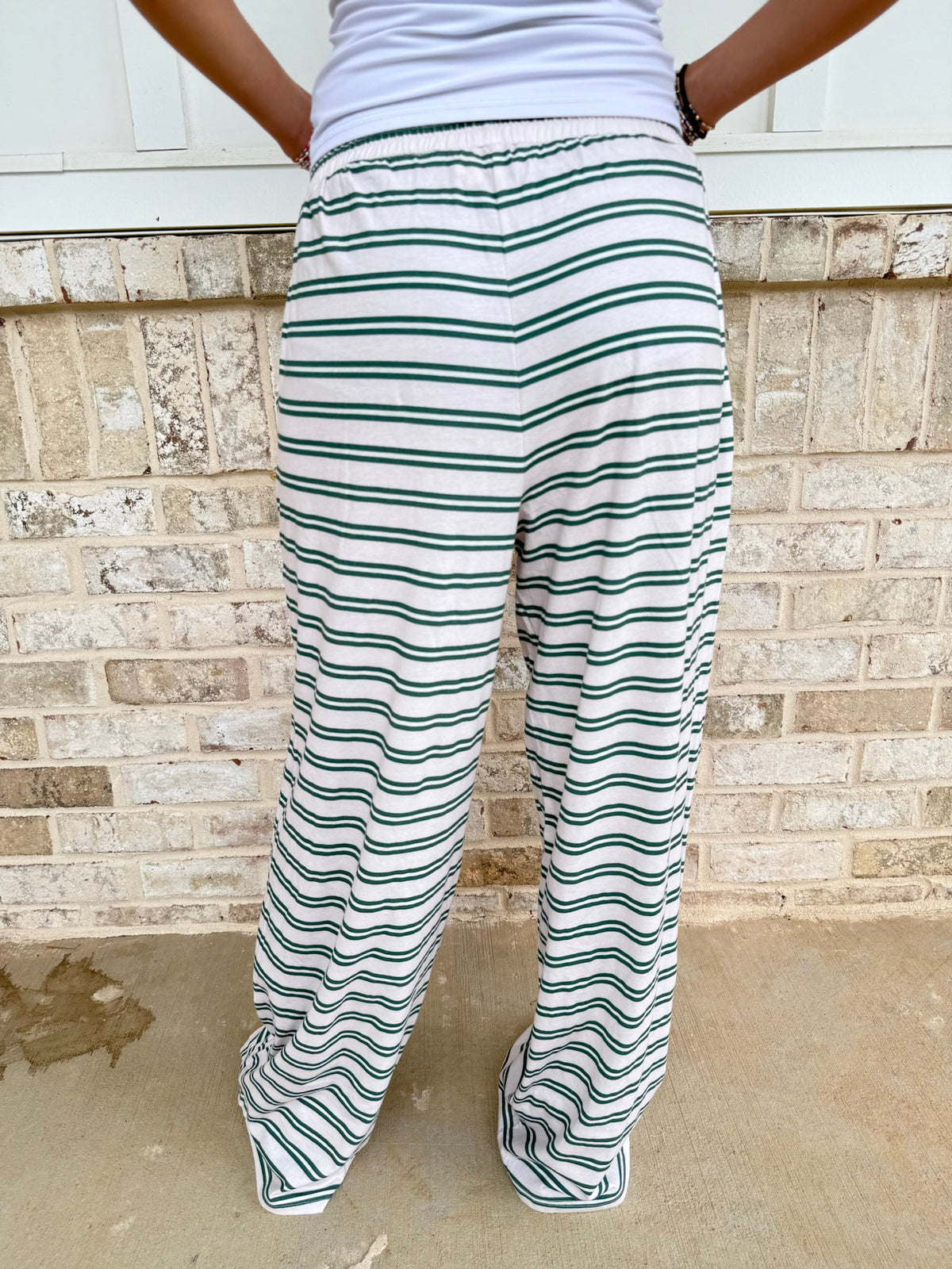 A person shown from the back wearing loose white pants with thin green horizontal stripes, standing outdoors against a light brick wall.