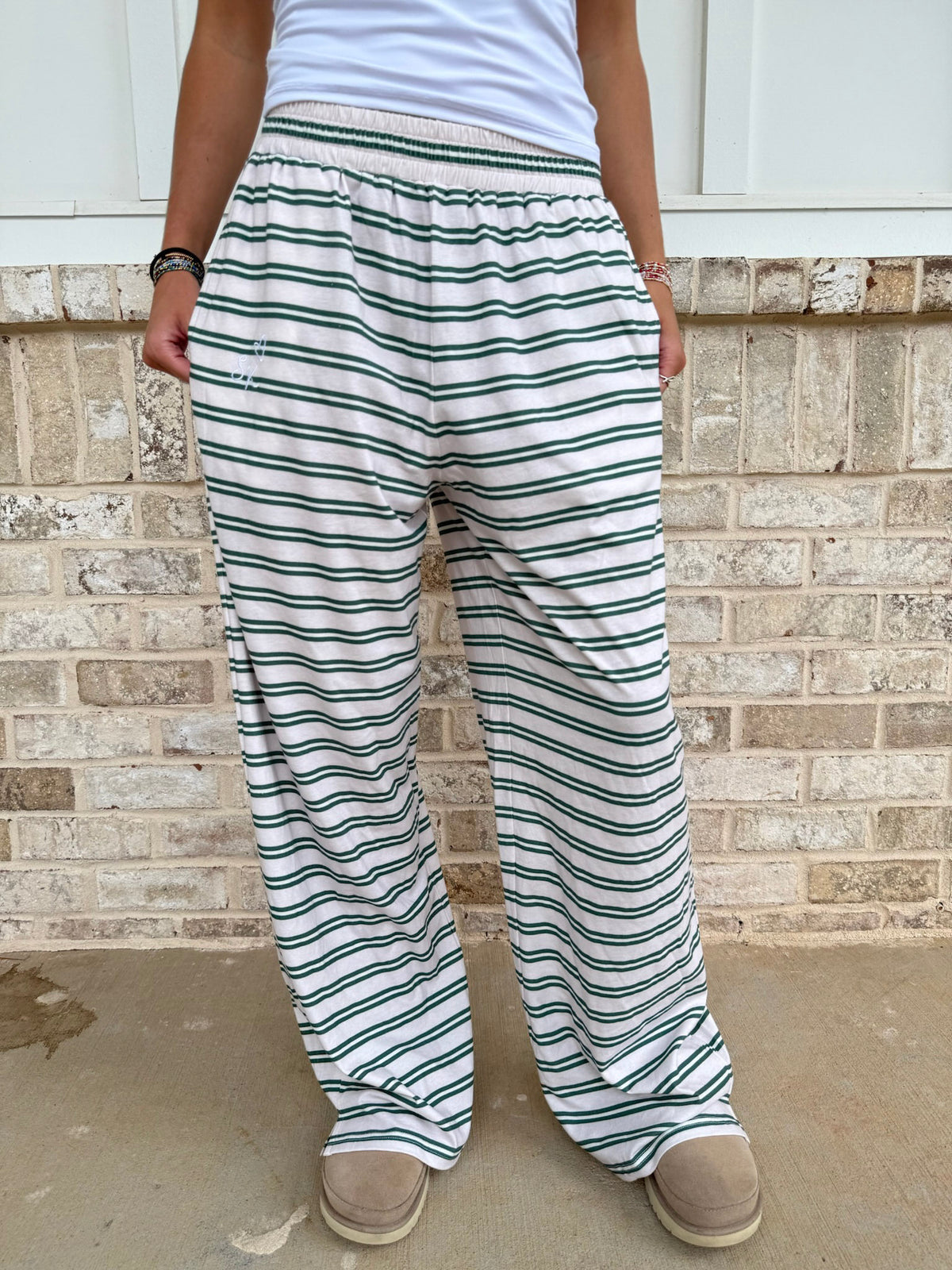 A person wearing loose, wide-leg striped pants with a white top and beige slippers, standing against a light brick wall.