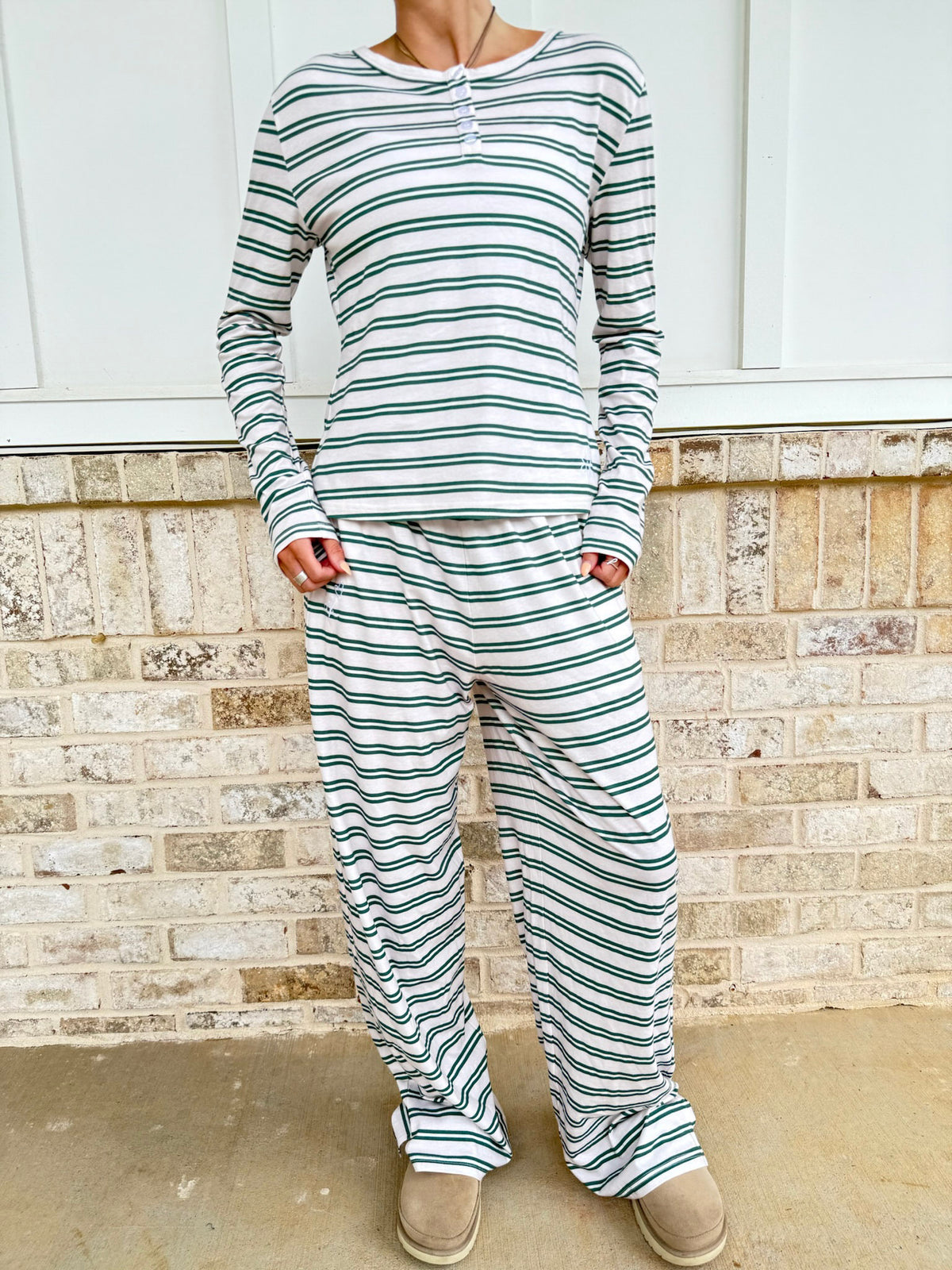A woman wearing a matching two-piece loungewear set with long-sleeve henley top and wide-leg pants, both featuring green and white horizontal stripes, standing in front of a brick wall.