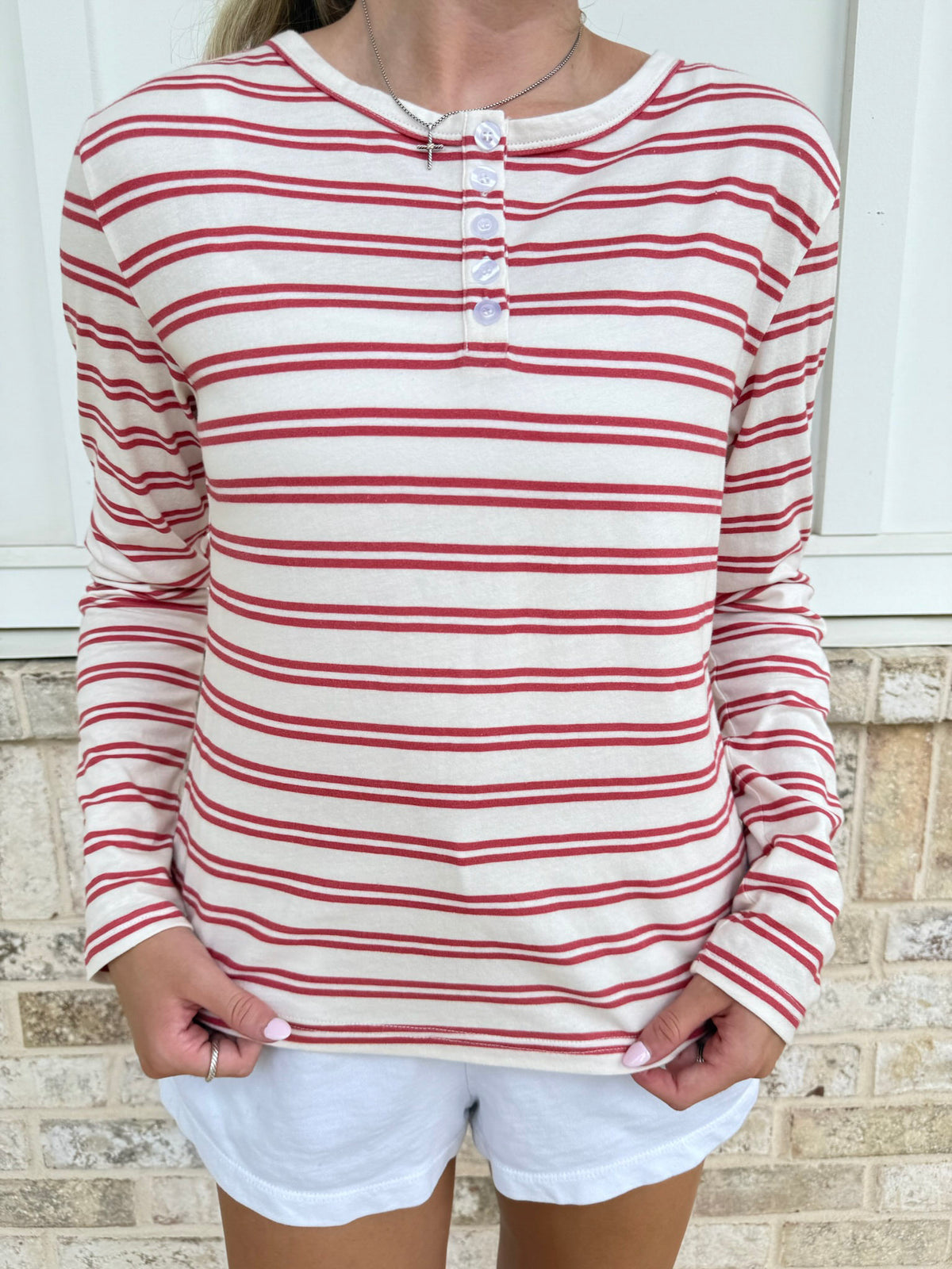A woman wearing a long-sleeve henley top with horizontal red and off-white stripes, paired with white shorts, standing in front of a light-colored brick wall.
