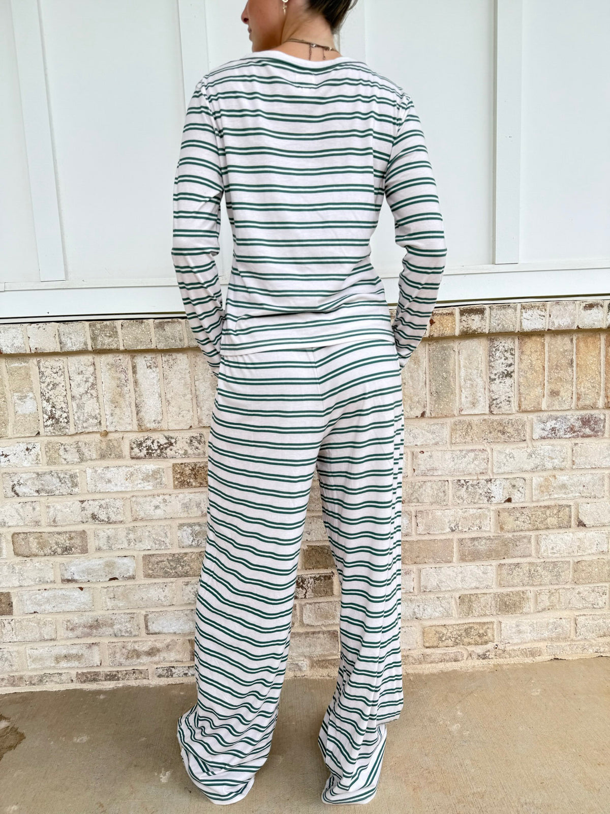 A person viewed from behind wearing a matching long-sleeve top and wide-leg pants set with thin dark green horizontal stripes on an off-white base, standing against a light brick wall.
