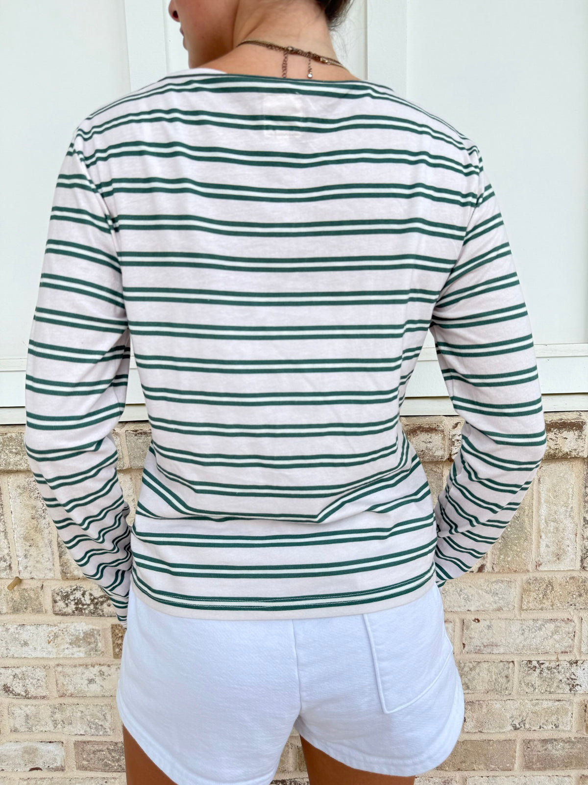 A person shown from the back wearing a long-sleeve striped top with horizontal green and white stripes, paired with white shorts, standing against a light brick wall.