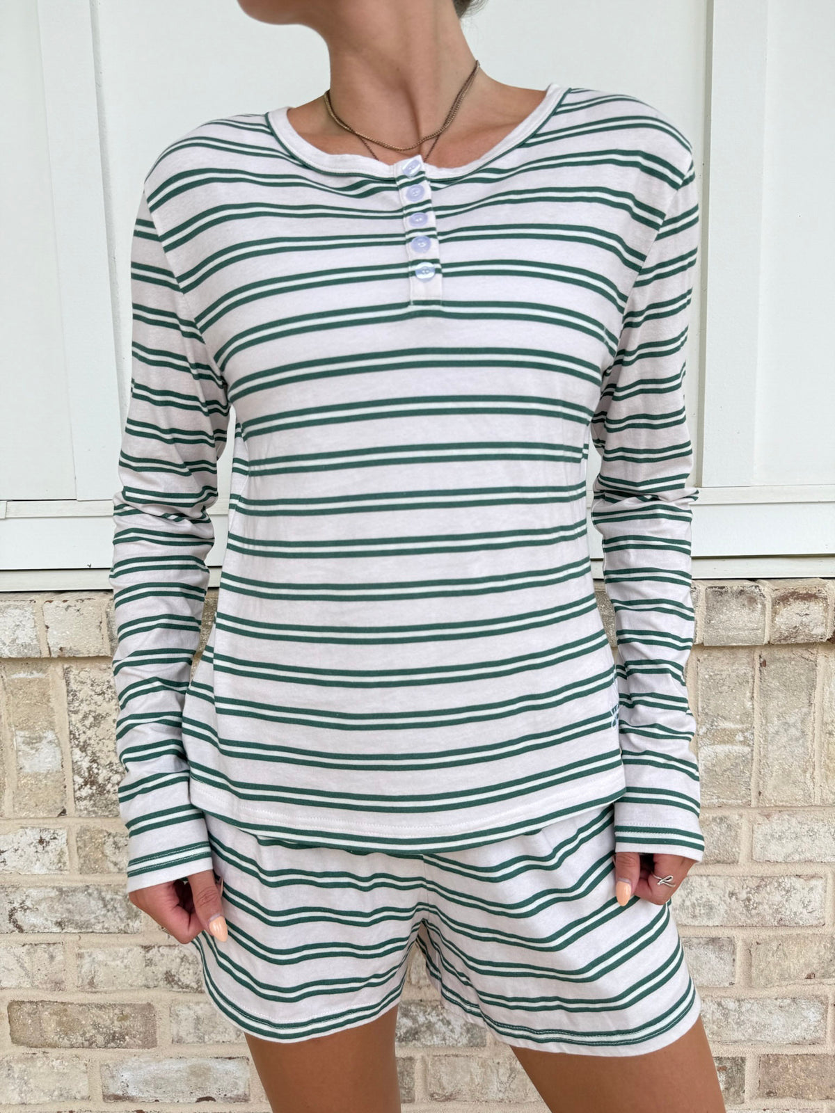A woman wearing a matching long-sleeve henley top and shorts set with horizontal green and white stripes, standing in front of a white paneled wall and light brick base.