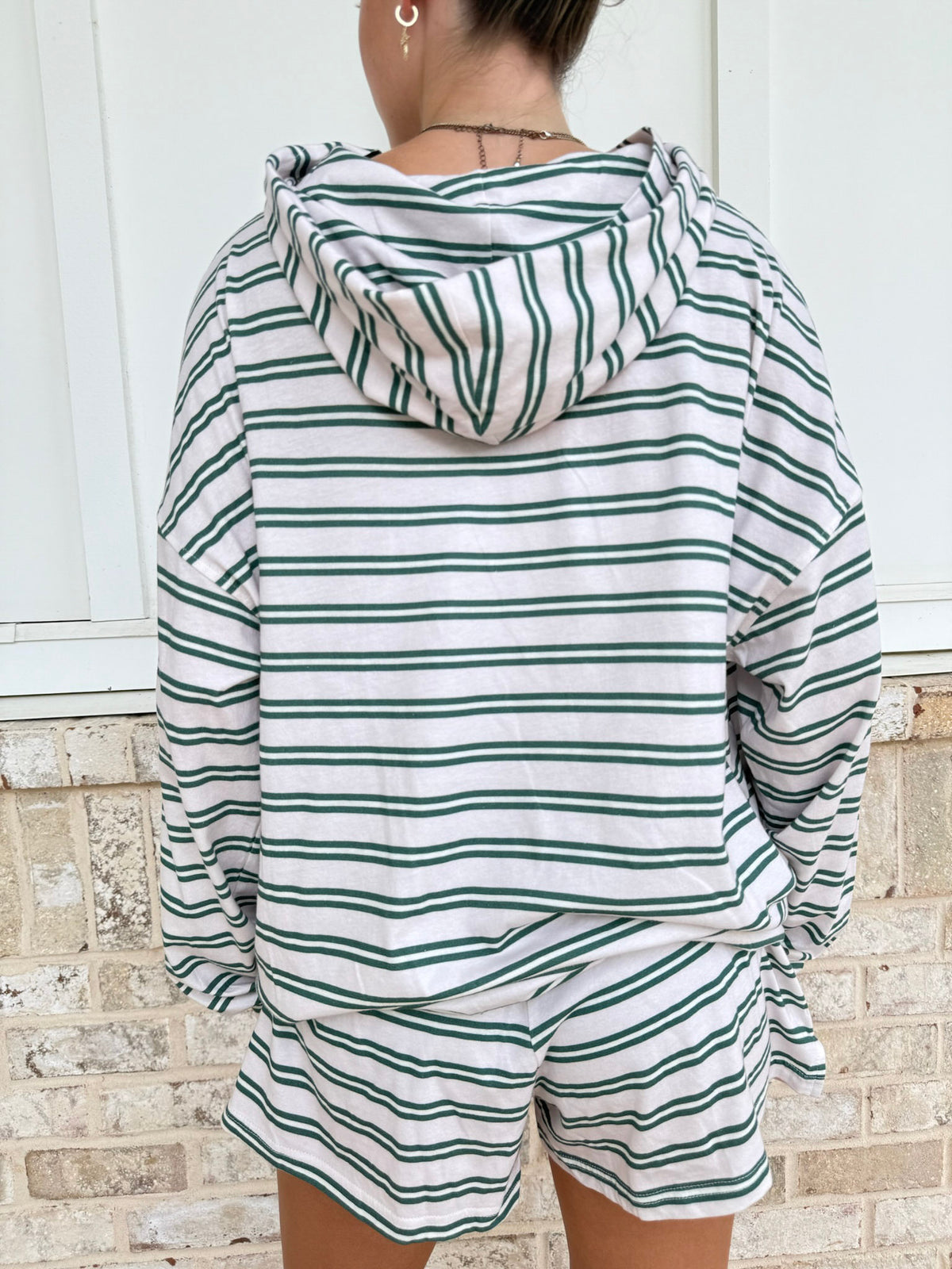 A person shown from the back wearing a matching green and white striped lounge set, featuring a hooded sweatshirt and shorts, set against a white paneled wall and light brick wall background.