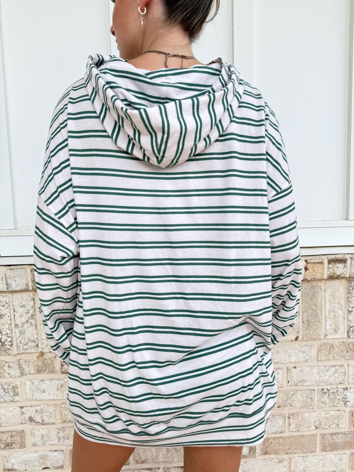 A person shown from the back wearing an oversized hooded sweatshirt with horizontal green and white stripes, standing against a white paneled wall and light brick base.