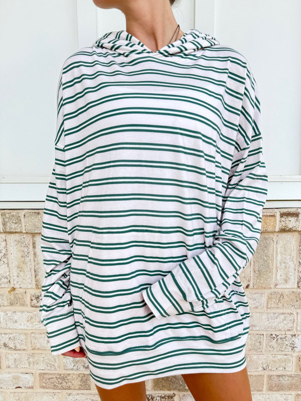 A person wearing an oversized long-sleeve hooded sweatshirt dress with horizontal green and white stripes, standing against a light brick wall.