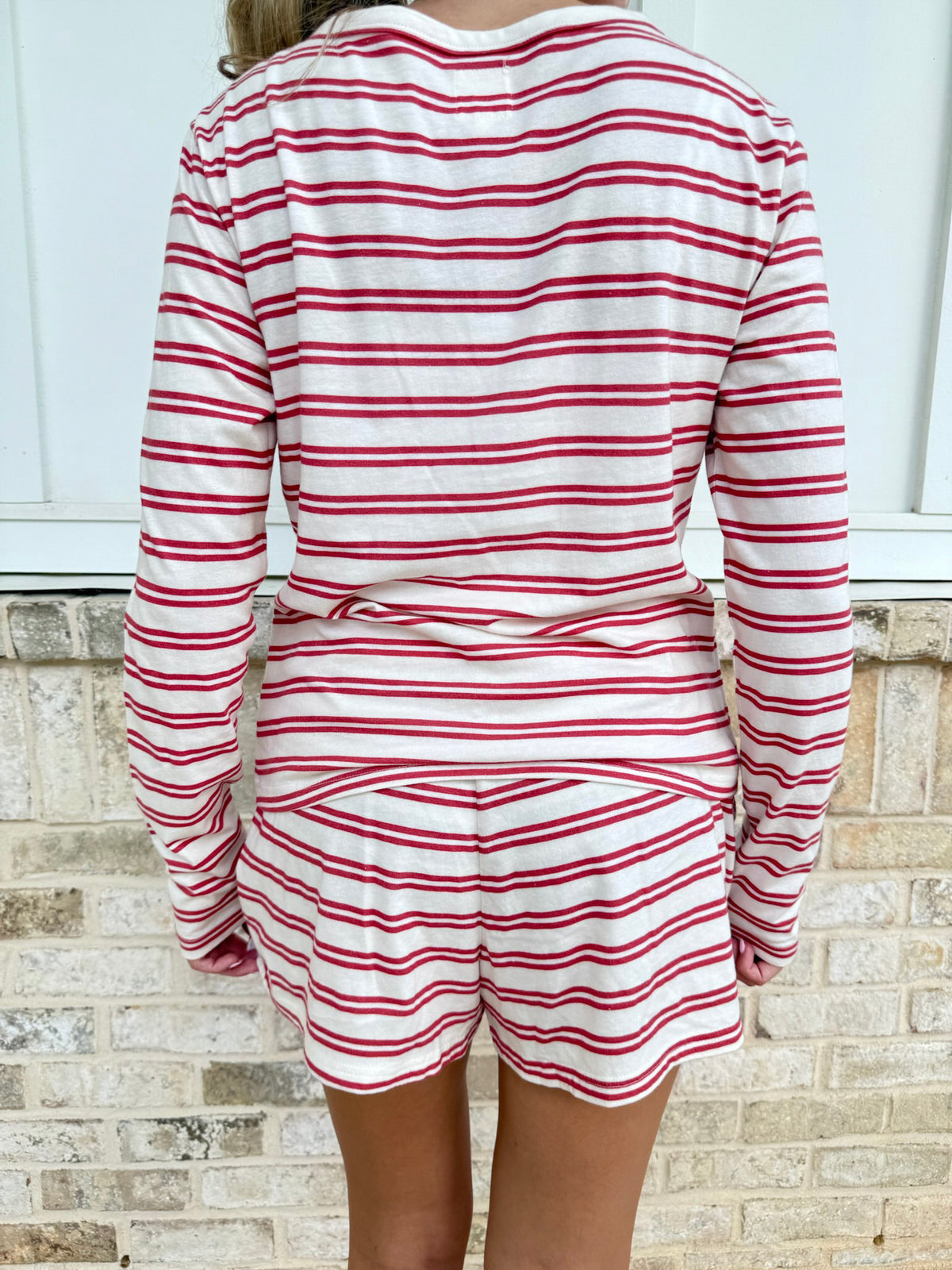 A person shown from the back wearing a matching long-sleeve top and shorts set with horizontal red and white stripes, standing against a light brick wall.