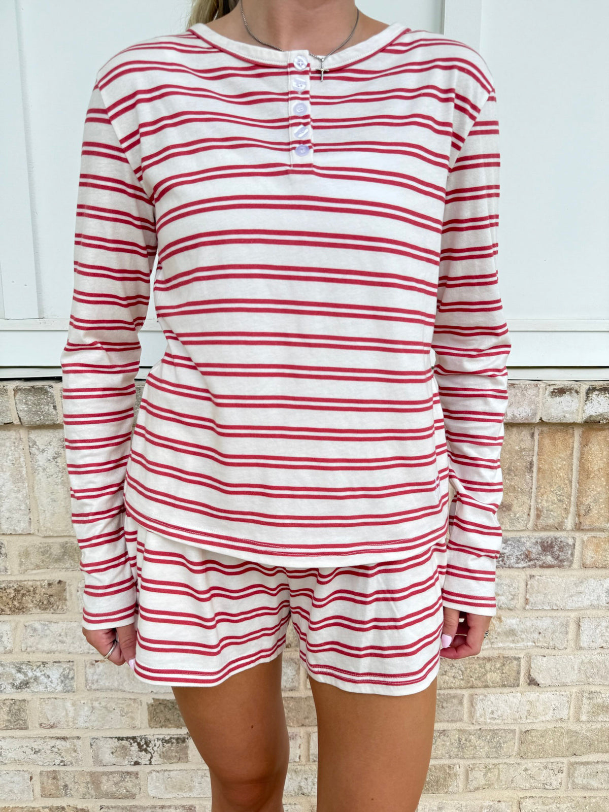 A woman wearing a matching long-sleeve henley shirt and shorts set with horizontal red and white stripes, standing in front of a light-colored brick wall.