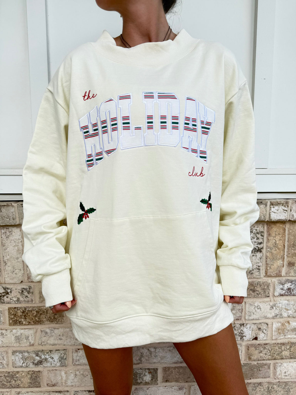 A person wearing an oversized cream sweatshirt with a large arched 'HOLIDAY' graphic outlined in red, with smaller red script text and small holly motifs near the front pockets, standing against a white door and brick wall.