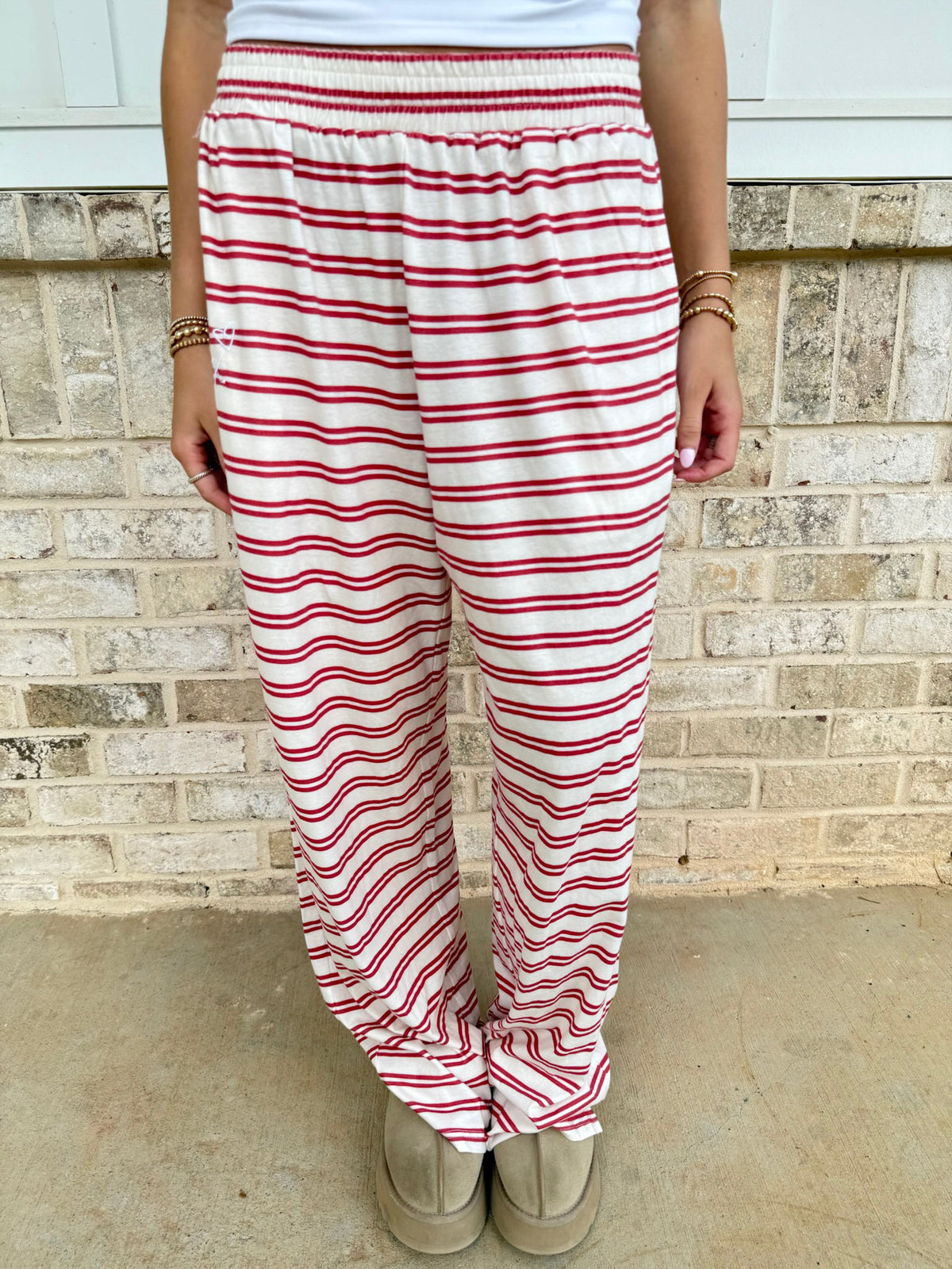 A person wearing loose white pants with thin red horizontal stripes, standing in front of a light beige brick wall.