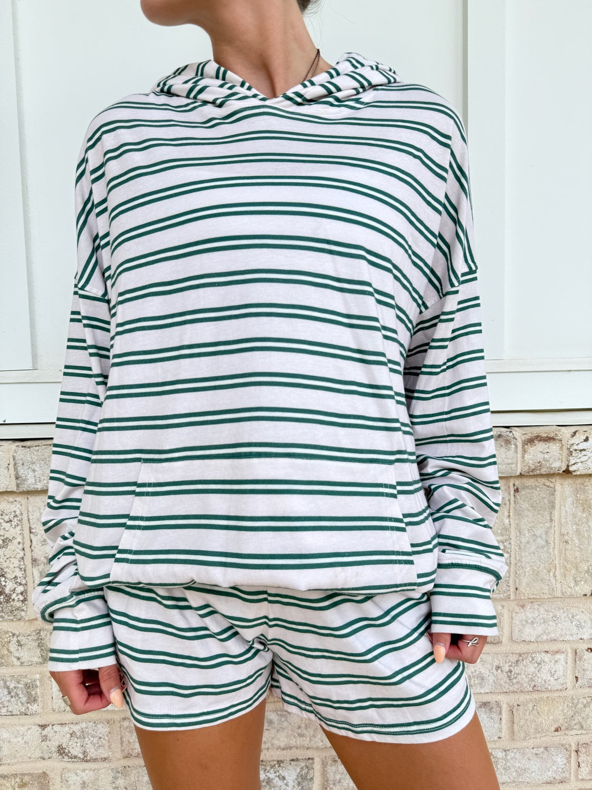 A person wearing a matching green and white striped lounge set, featuring a long-sleeve hooded sweatshirt and coordinating shorts, standing against a white paneled wall and light brick base.
