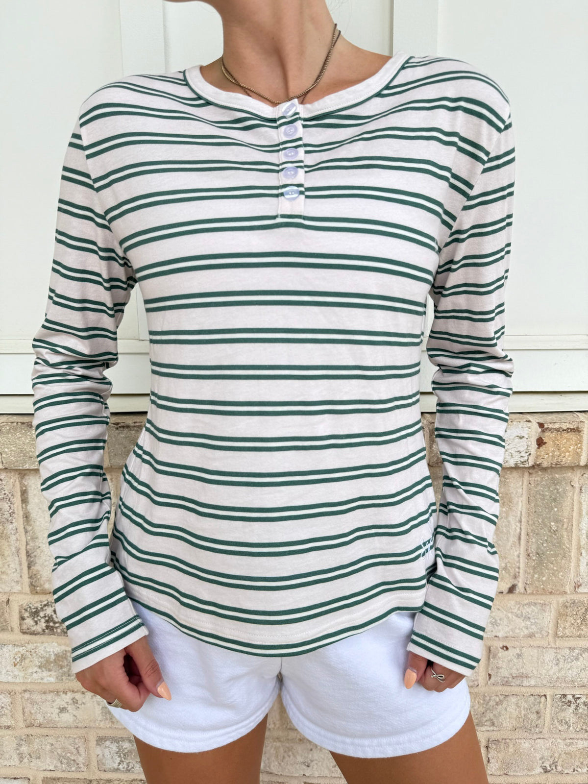 A woman wearing a long-sleeve henley top with horizontal green and white stripes, paired with white shorts, standing against a white door and light brick wall.