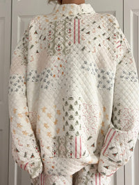 A person wearing a quilted long-sleeve top and matching pants in an off-white patchwork fabric with a multicolor holiday print of pine trees, pinecones, and geometric patterns.