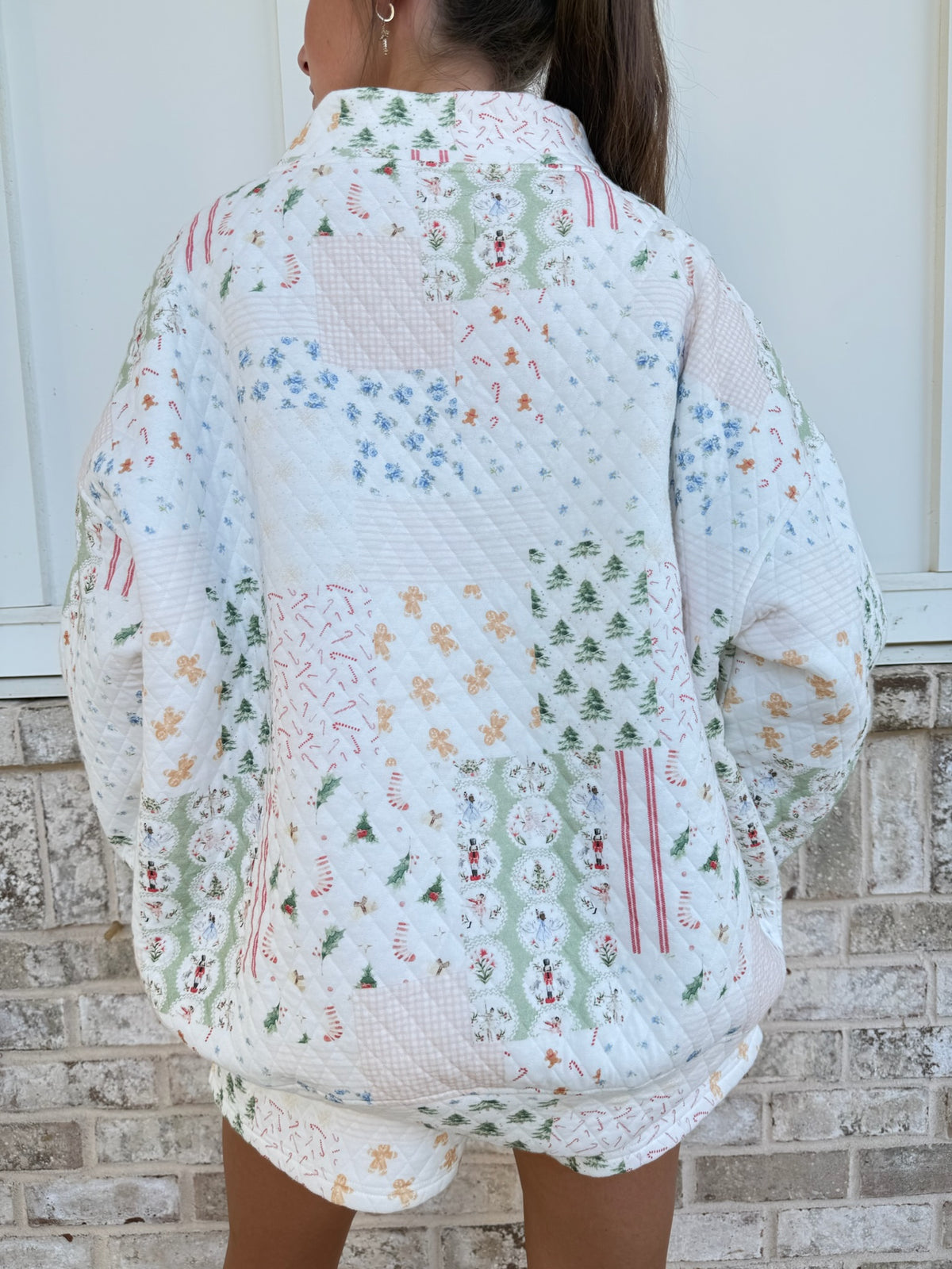 A person viewed from behind wearing a quilted white patchwork jacket with colorful Christmas-themed prints, standing in front of a light-colored brick wall.