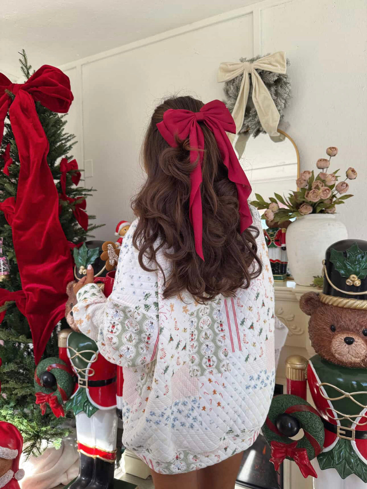 A woman viewed from behind wearing a white knit sweater with colorful holiday patterns and a large dark pink hair bow, standing in a decorated Christmas room with nutcrackers and a tree.