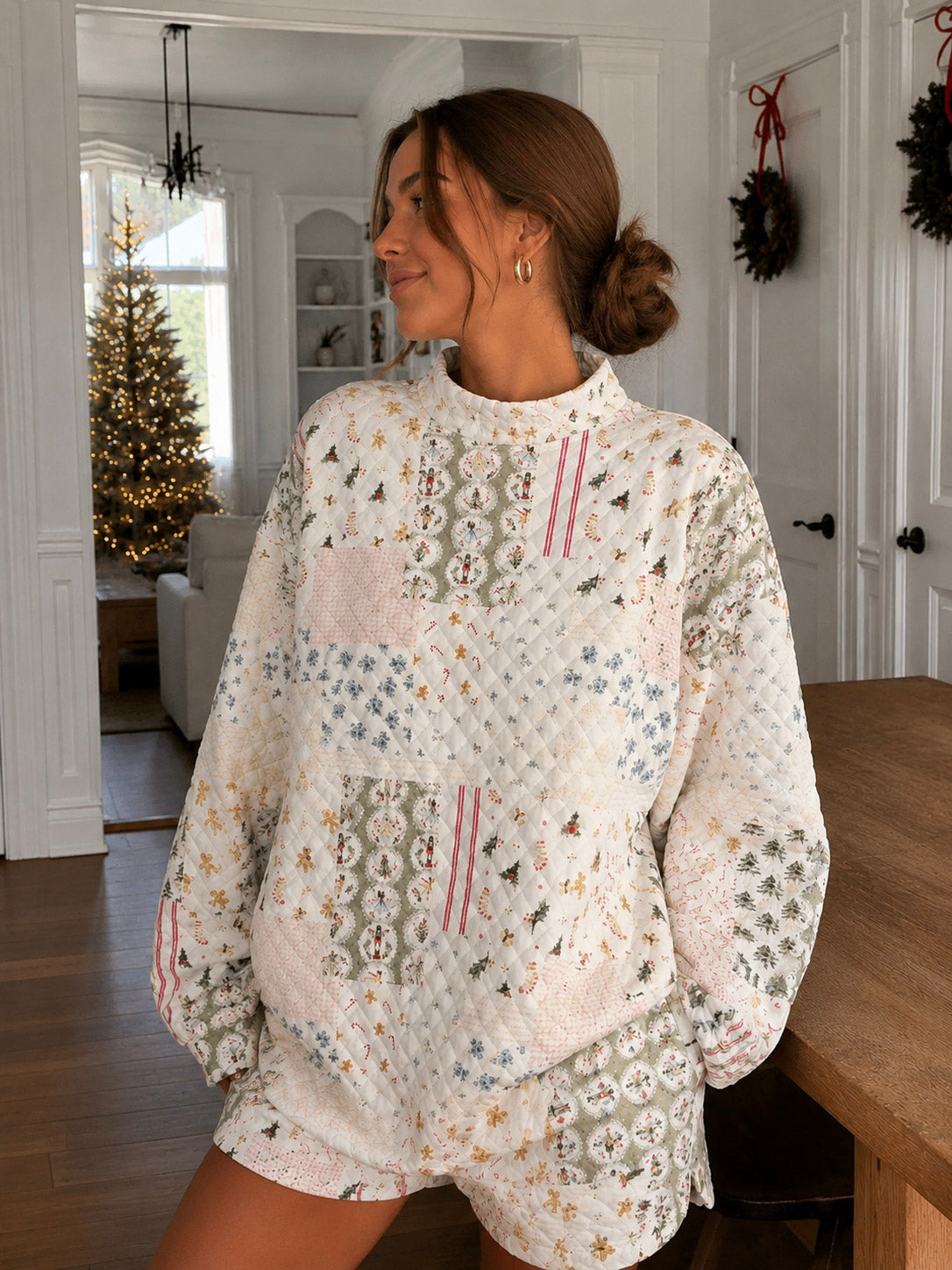 A woman wearing a cozy, oversized quilted patchwork pajama set with a Christmas tree and snowflake pattern, standing in a bright, holiday-themed living room.