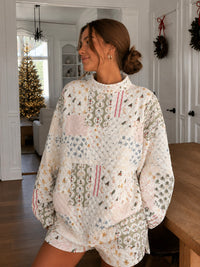 A woman wearing a cozy, oversized quilted patchwork pajama set with a Christmas tree and snowflake pattern, standing in a bright, holiday-themed living room.