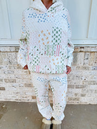 A person wearing a matching quilted white hoodie and jogger pants set with a multicolor Christmas patchwork pattern, standing in front of a brick wall.