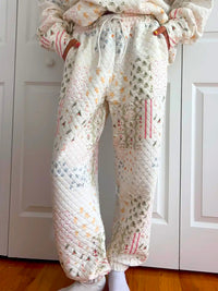 A person wearing quilted jogger-style pants with a patchwork pattern in soft pastel colors, standing in front of white paneled closet doors and a wood floor.