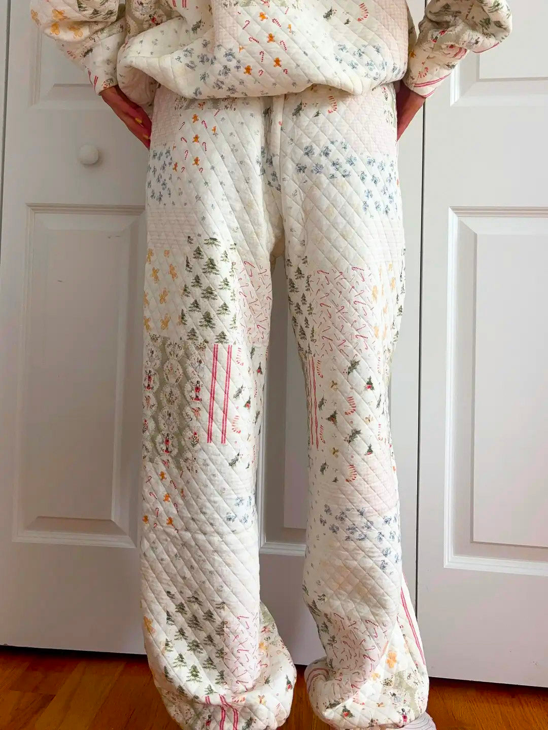 A person wearing quilted pajama pants with a patchwork design of small floral and tree prints in pastel colors, standing in front of white paneled doors.