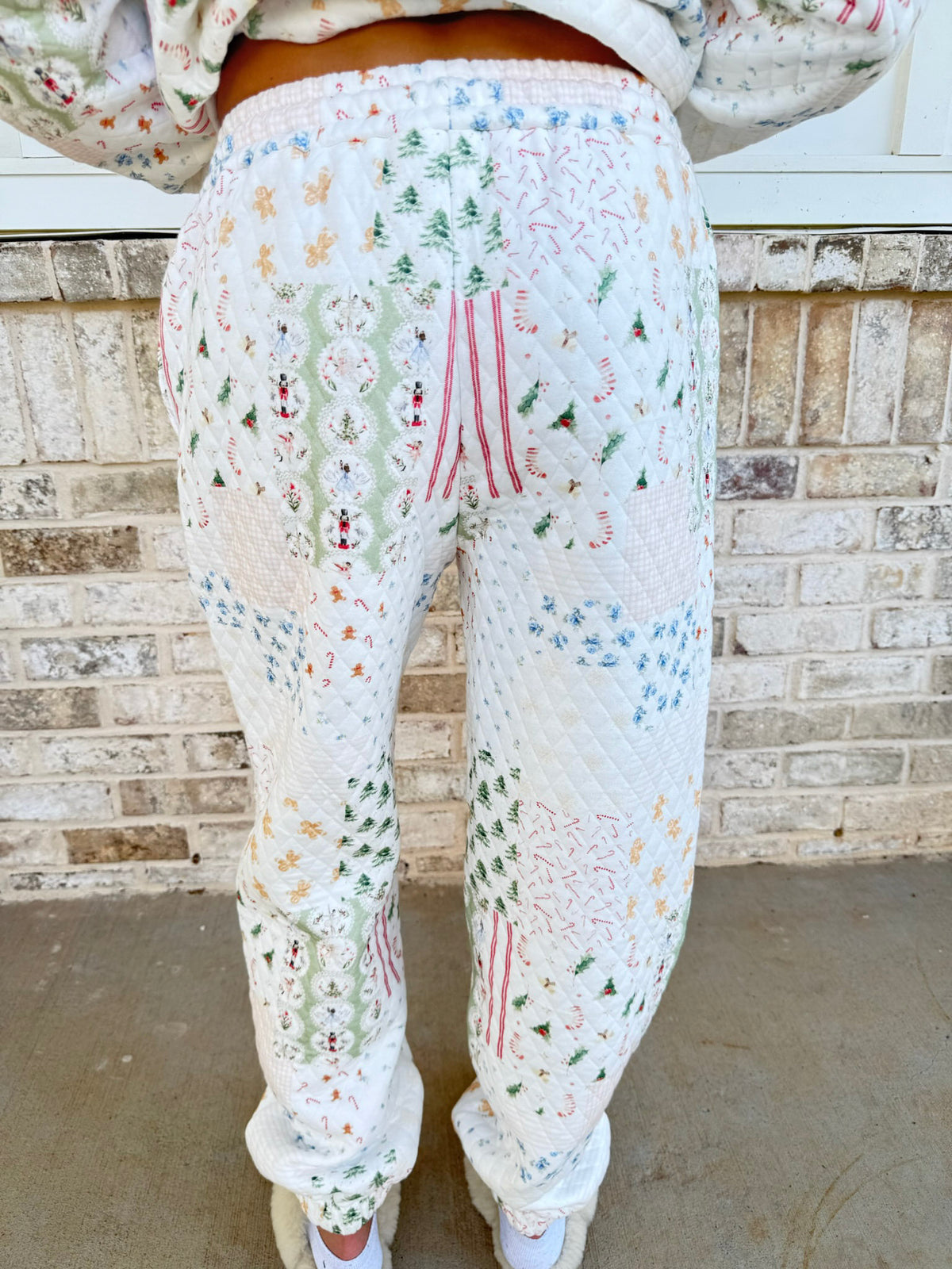 A person viewed from behind wearing quilted white pants with a multicolor Christmas-themed patchwork pattern of trees, ornaments, and snowflakes, standing in front of a brick wall.