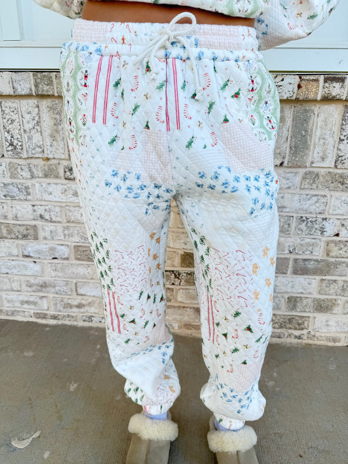 A person wearing quilted white jogger-style pants with a multicolor Christmas print, standing in front of a brick wall.