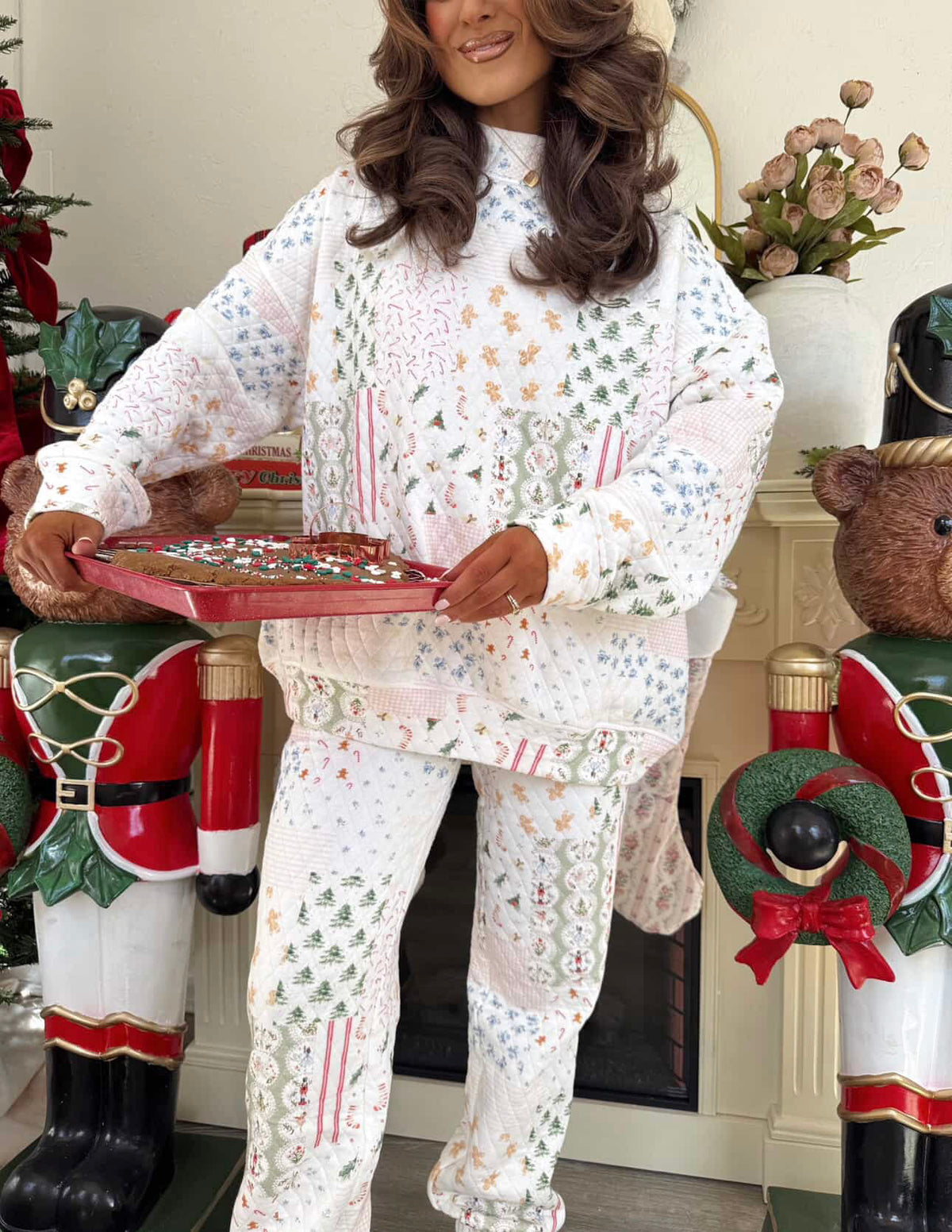 A woman wearing a festive Christmas-themed pajama set with a patchwork print of candy canes, snow globes, and evergreen trees, holding a red tray of treats in a holiday decorated room.