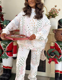 A woman wearing a festive Christmas-themed pajama set with a patchwork print of candy canes, snow globes, and evergreen trees, holding a red tray of treats in a holiday decorated room.