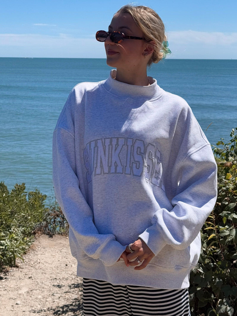 Person wearing a white sweatshirt with 'sunkissed' text, standing by the ocean.