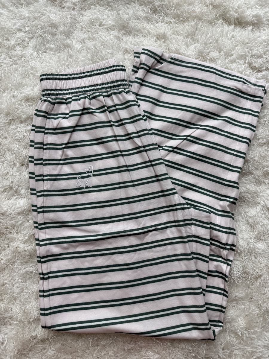 A pair of folded striped lounge pants with an elastic waistband, featuring horizontal stripes in dark green and black on a light pinkish-beige base, laid flat on a white shag rug.