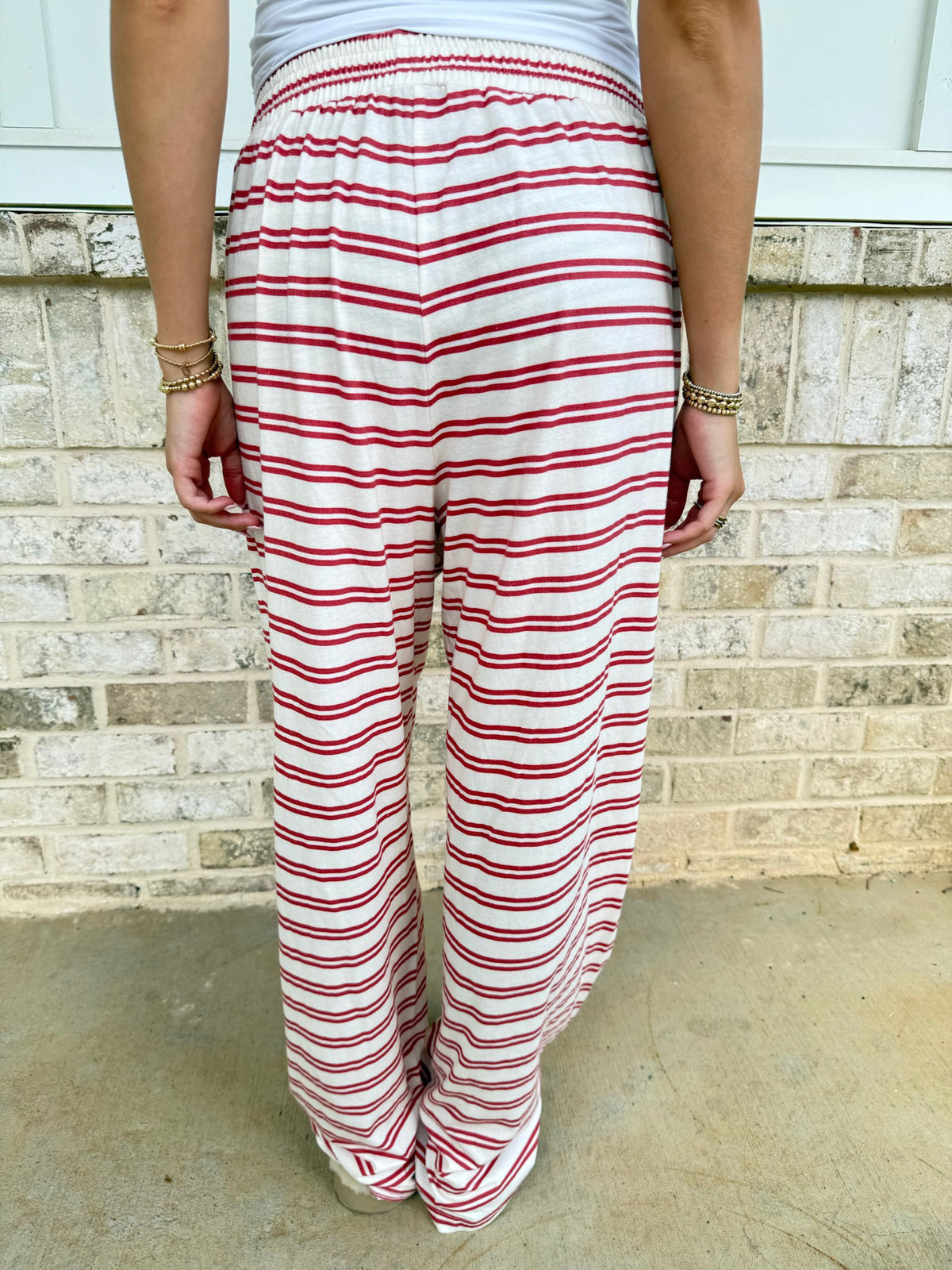 A person shown from the back wearing loose, wide-leg pants with red and white horizontal stripes, standing against a light brick wall.