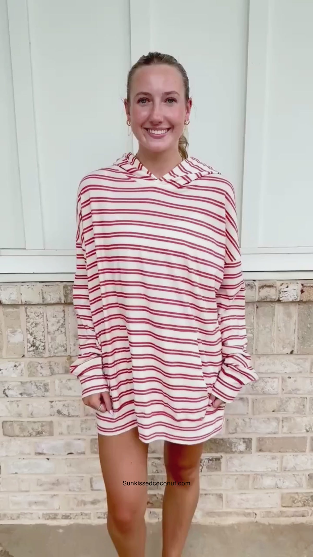 A person wearing a matching red and white striped lounge set with a hooded sweatshirt and sweatpants, standing in front of a light brick wall.