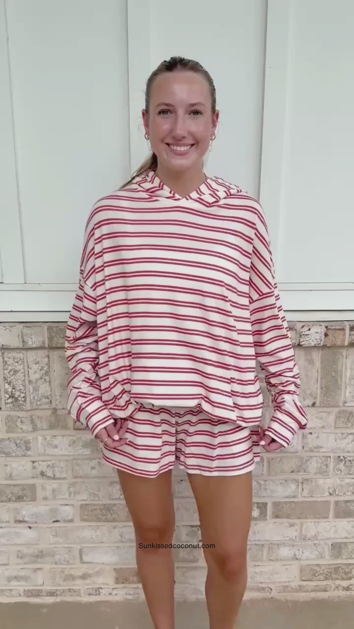 A person wearing a matching red and white striped lounge set with a hooded sweatshirt and sweatpants, standing in front of a light brick wall.