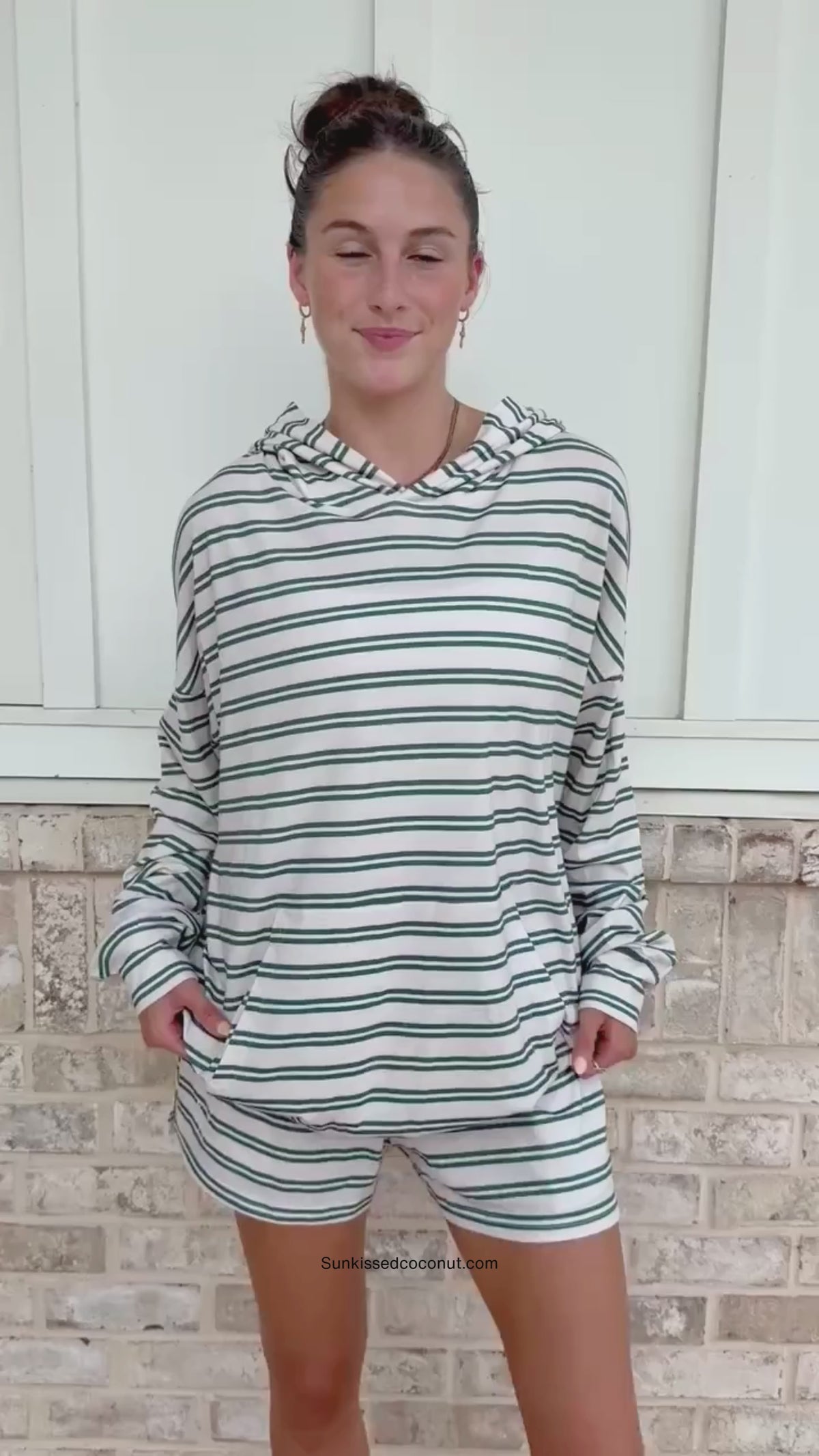 A person wearing a matching green and white striped lounge set with a hooded sweatshirt and sweatpants, standing in front of a light brick wall.