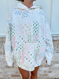 A person wearing an oversized patchwork quilted hoodie with a white base and multicolor floral, tree, and stripe patterns, standing against a light brick wall.