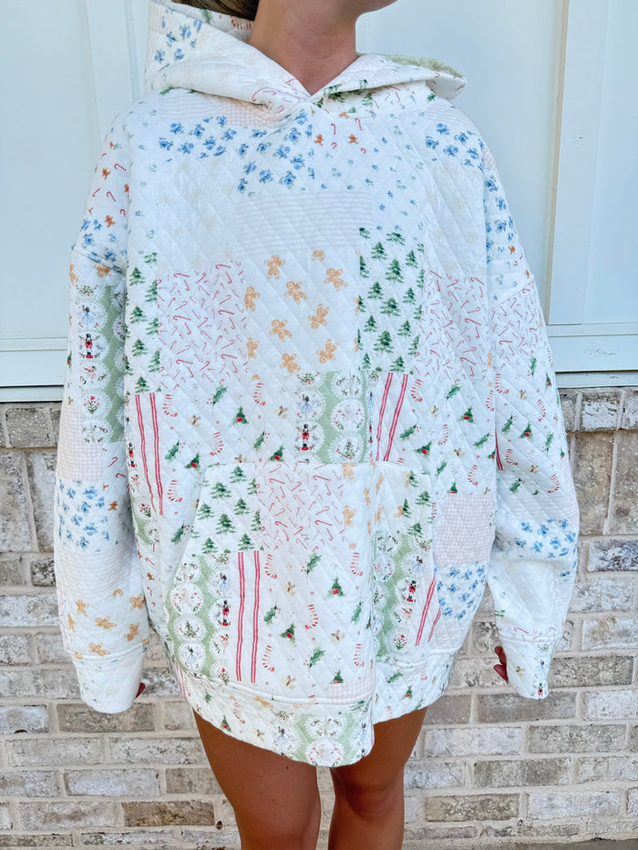 A person wearing an oversized patchwork quilted hoodie with a white base and multicolor floral, tree, and stripe patterns, standing against a light brick wall.