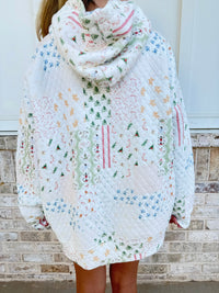 A person wearing a quilted, oversized hooded sweater with a multicolor Christmas patchwork pattern, standing in front of a light brick wall.