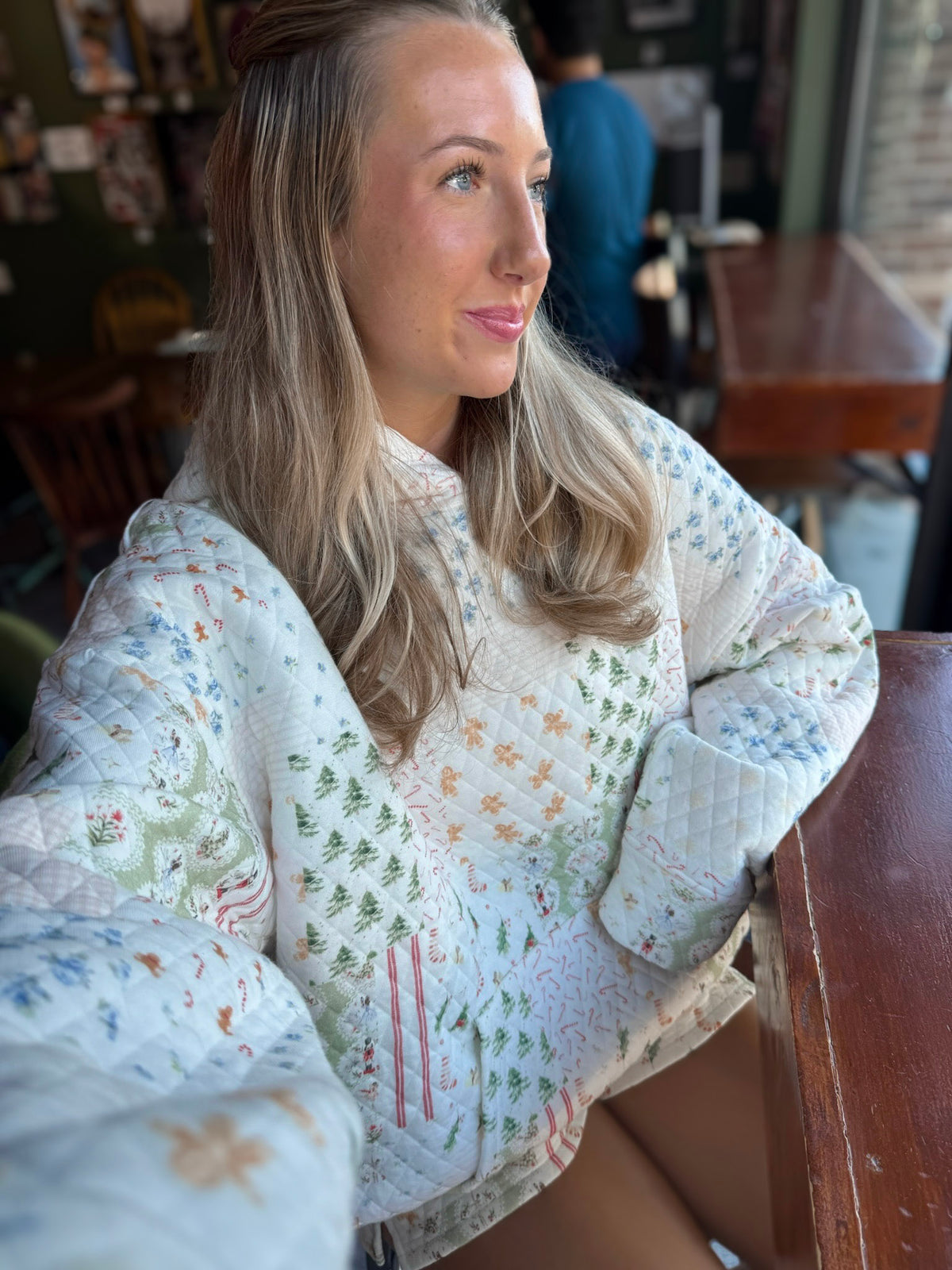 A woman wearing a quilted white jacket with a colorful Christmas tree and holiday pattern, sitting in a cozy indoor setting.