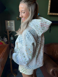 A woman wearing a quilted hooded jacket with a multicolor floral pattern, photographed in a cozy indoor setting with a dark green wall and brown leather chair in the background.