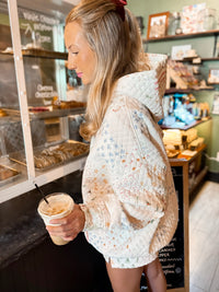 A woman in a cozy quilted hoodie with a floral and star pattern stands in a cafe, holding an iced coffee. The hoodie features a cream base with light blue, pink, and orange floral details and small star prints.