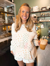 A smiling blonde woman stands inside a cafe holding an iced coffee, wearing a white knit matching top and shorts set with a multicolor holiday tree pattern.